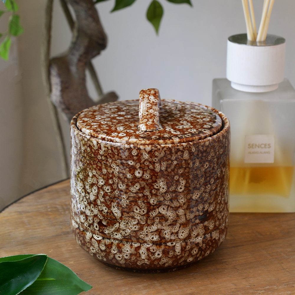 Decorative ceramic jar with a textured brown and white pattern on a wooden surface, with a bottle of perfume in the background.
